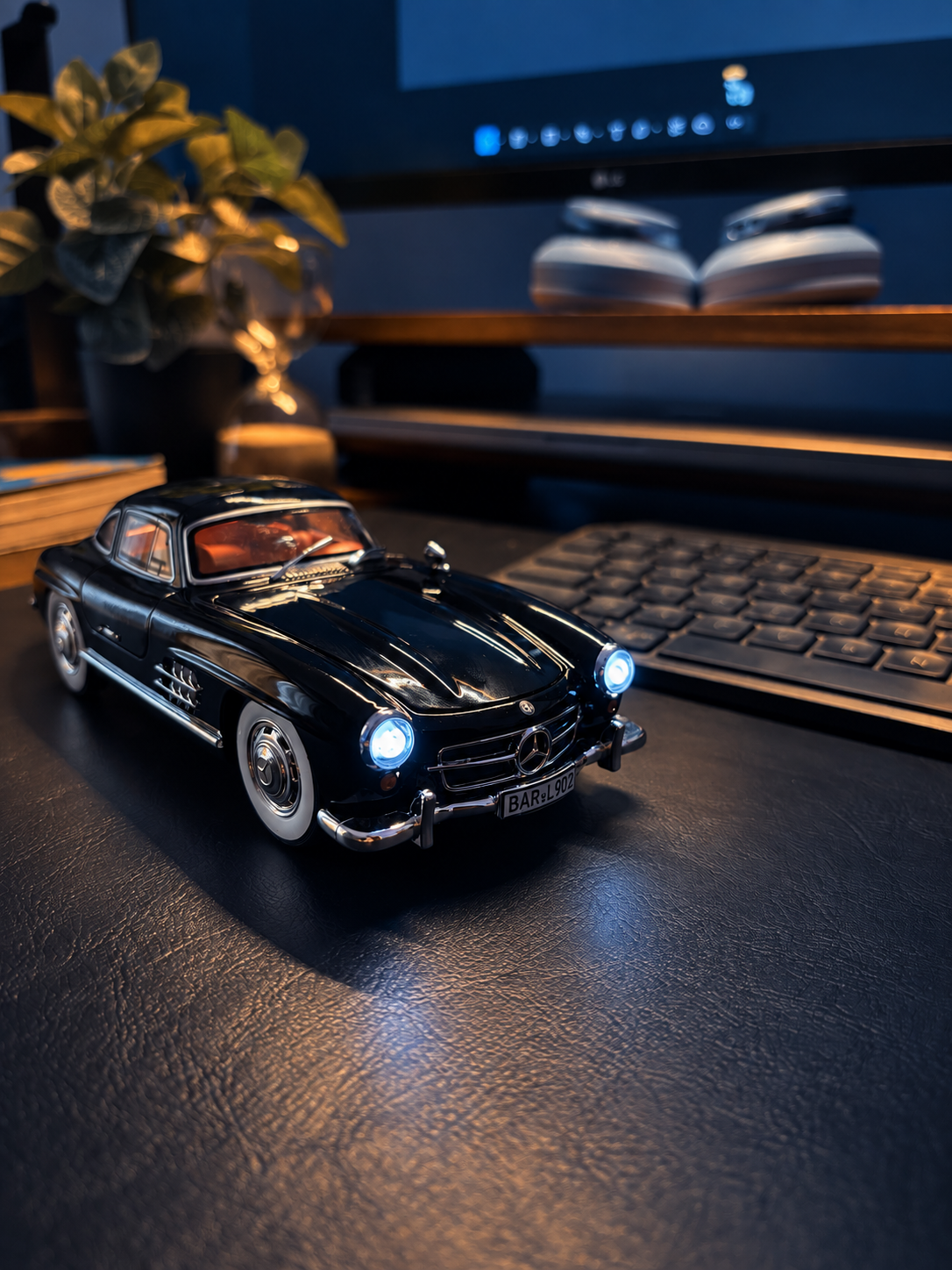 A black model Mercedes with glowing headlights on a desk, with a keyboard, monitor, headphones, and plant behind it